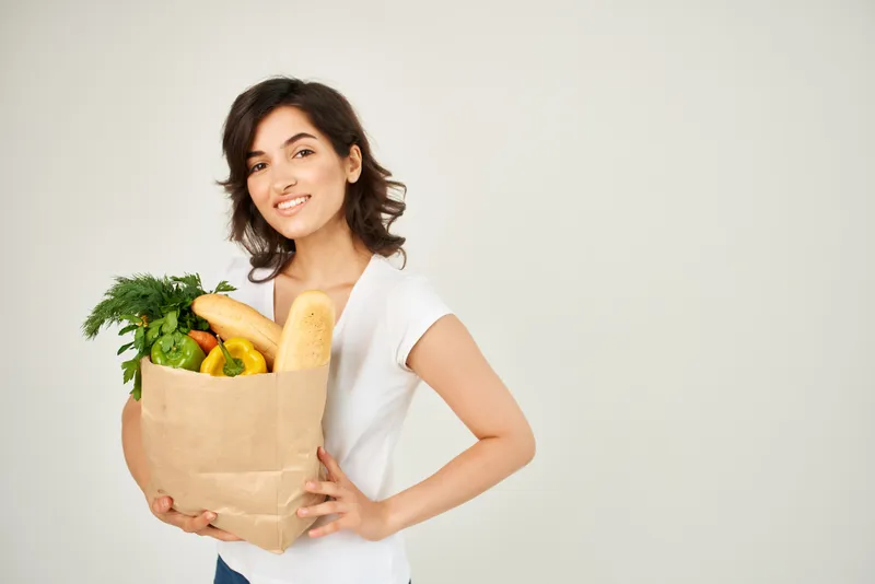 Весёлая женщина с пакетом продуктов из супермаркета на светлом фоне
