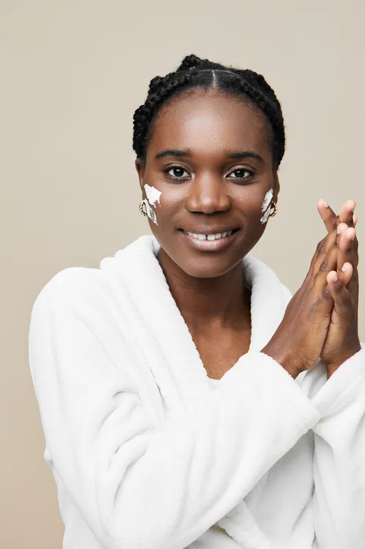 A joyful Black woman applying skincare in a white robe, showcasing a healthy skincare routine for glowing complexion, set against a soft beige background