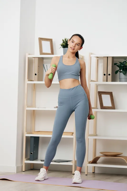 Woman fitness enthusiast in athletic gray outfit stands in a bright home gym, lifting dumbbells with controlled form for a focused strength training session.