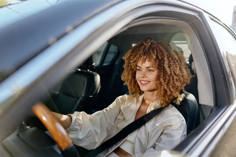 Smiling woman driving car with curly hair wearing light blouse, enjoying sunny day, modern interior with leather seats, safety belt fastened.