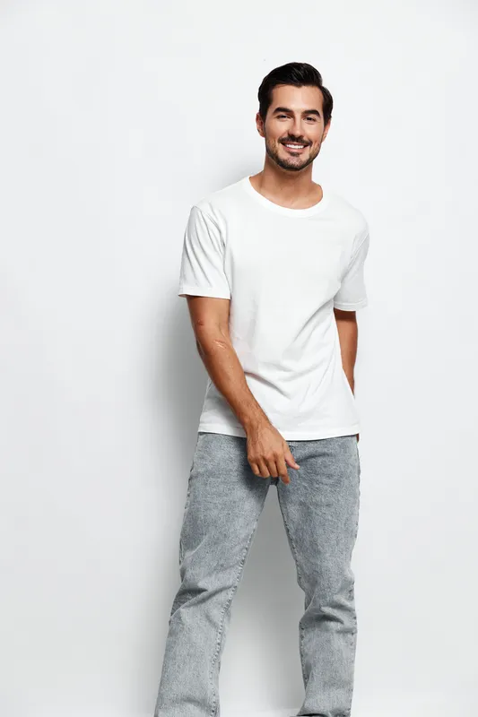Full body smiling happy cheerful young caucasian man wearing white t-shirt and gray pants posing confidently against plain solid background in studio portrait. People lifestyle concept.
