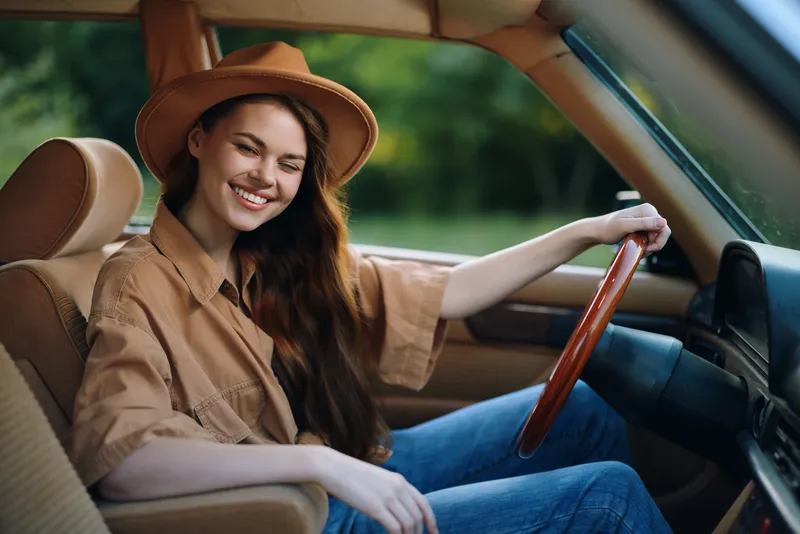 Smiling young woman with long hair wearing a hat driving a vintage car in nature, conveying joy and freedom during summer road trip.