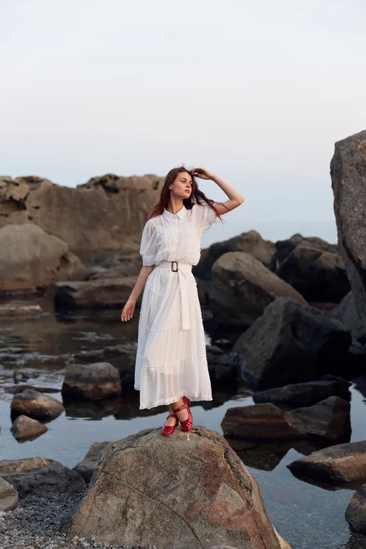 Безмятежная женщина в белом платье стоит на каменистом берегу, запустив руки в волосы и глядя на океан
