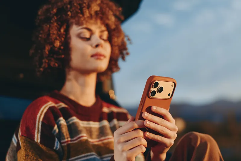 Woman lifestyle portrait outdoors, curly haired woman in red striped sweater holds smartphone, looks at screen, relaxed candid moment, sunset light, mountains blurred background, natural air