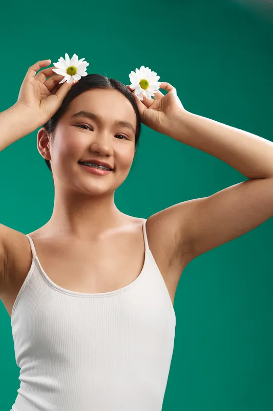Asian woman wearing a white tank top holds daisies above her head, smiling, in a vibrant studio scene that conveys freshness, playfulness, and joy, inviting a positive, modern lifestyle vibe.