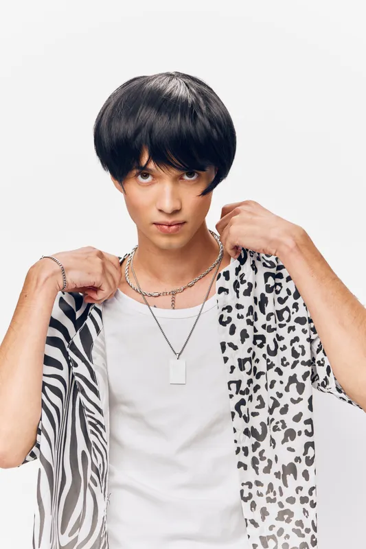 Young man with short dark hair wearing a white tank top and holding a black and white patterned shirt, looking directly with a neutral expression on a white background.