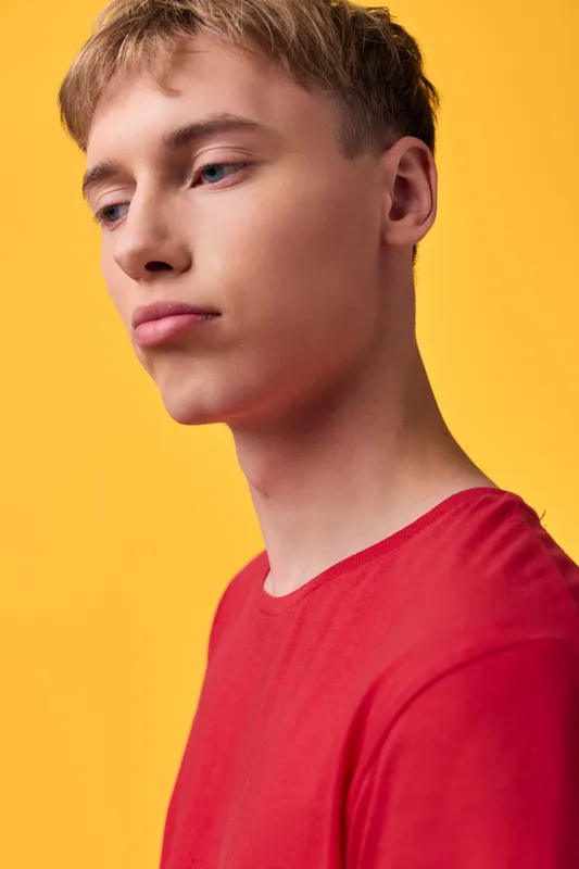 Young man wearing a red shirt against a vibrant yellow background, showcasing a minimal studio style with soft lighting, thoughtful expression, fashion and lifestyle mood