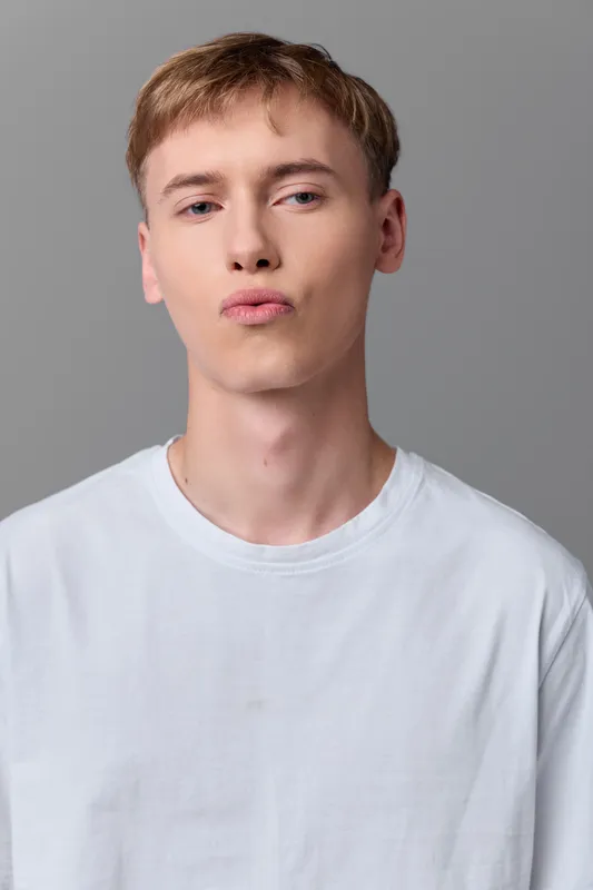 A young man with a pout and lip expression wears a white tee in a studio shot against a neutral background, captured in a head and shoulders frame with clean, minimalist styling.