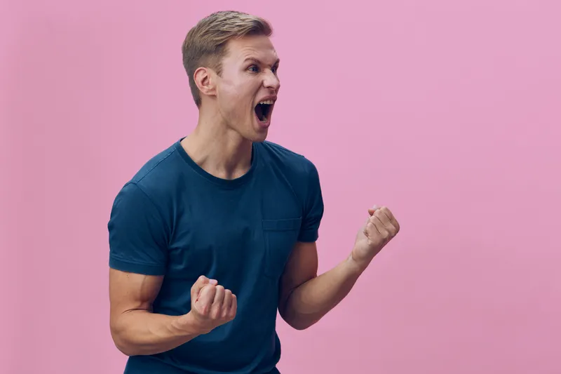 Young Caucasian man wearing a blue t-shirt expressing excitement with clenched fists and open mouth, isolated on a plain pink background, studio portrait, emotional reaction, people lifestyle concept.
