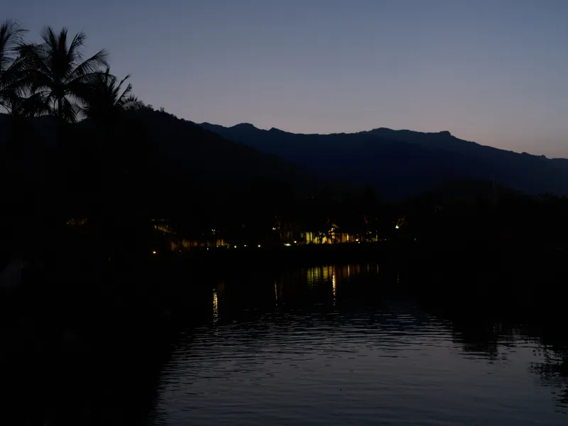Night river landscape with mountain silhouettes and palm trees reflecting city lights in calm water under dark sky.