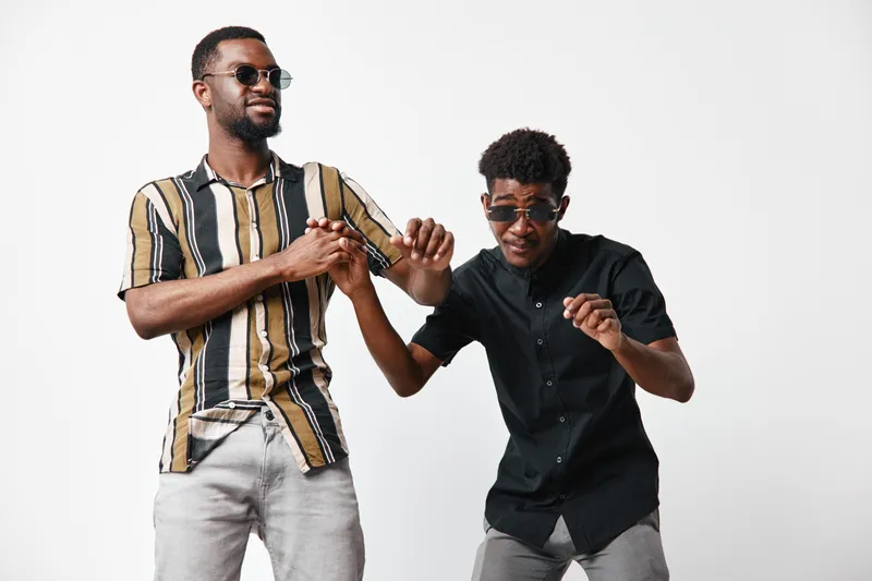 Two young black men with dark skin wearing sunglasses and casual shirts dancing and having fun together on white background in studio. Friendship and joy concept.