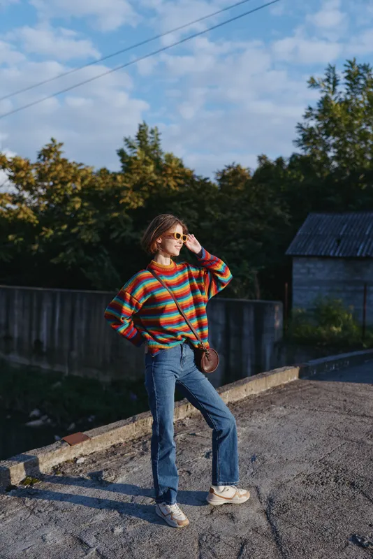 Young woman in a striped sweater and sunglasses wearing jeans, standing on a suburban street in sunlight. Casual outfit, confident pose with hand on hip and crossbody bag.