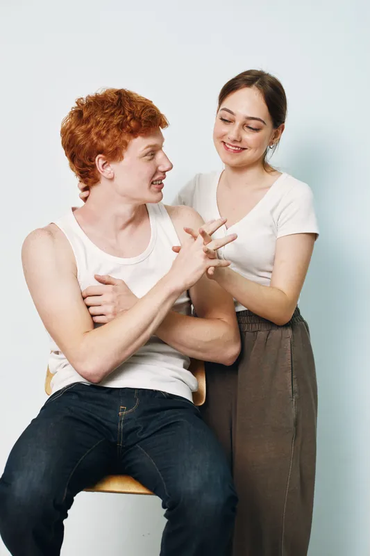 Young man with red hair sitting on chair smiling and interacting with woman standing nearby in casual white tops and relaxed atmosphere indoors bright background.