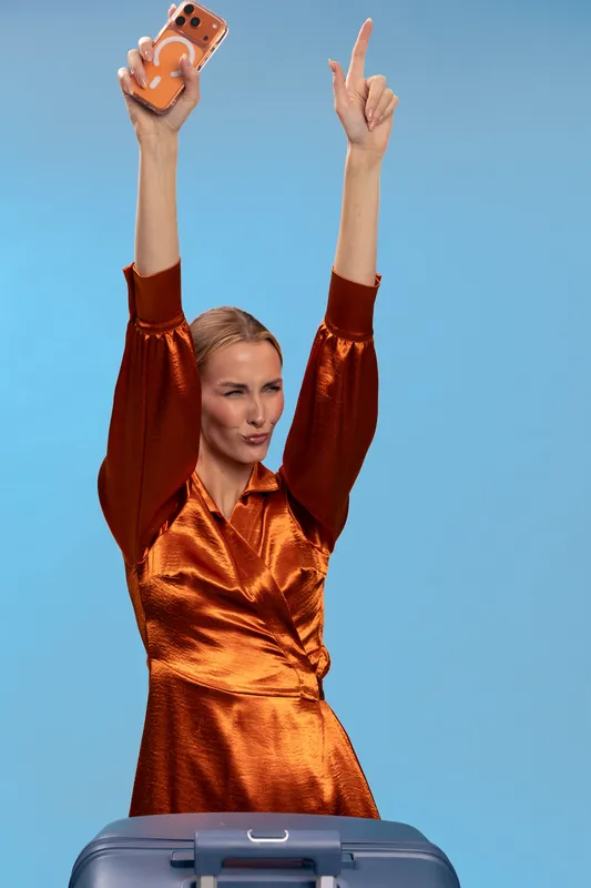 Happy woman in shiny orange dress holds phone up and gestures with finger, standing behind suitcase on blue background. Concept of travel excitement, vacation, and joyful celebration.