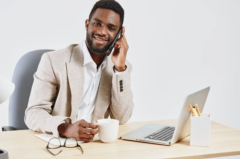 Professional smiling man of African descent in business casual attire working at desk, talking on mobile phone, with laptop and coffee cup, in modern office with light background, concept: workplace c