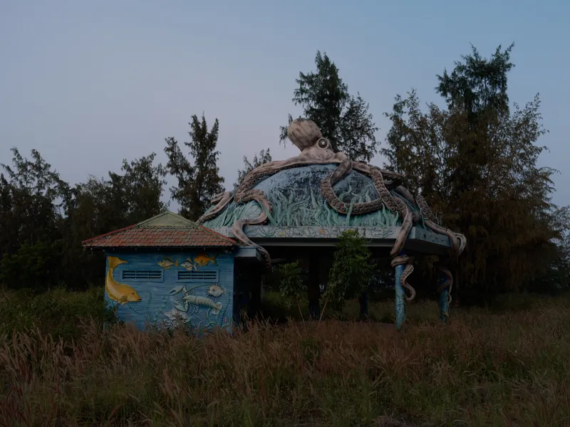 Abandoned building with large octopus sculpture on the roof, surrounded by tall grass and trees, captured during dusk with a moody sky in the background.