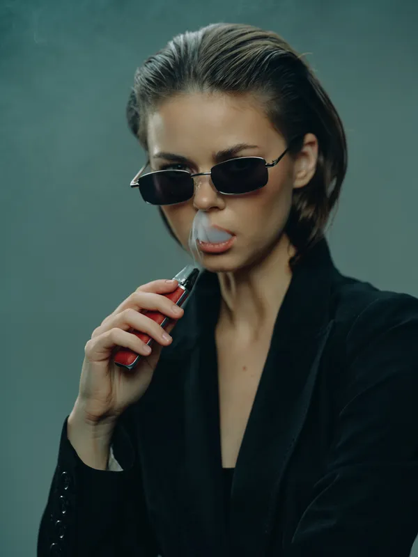 Young woman wearing sunglasses vaping in dark stylish outfit exhaling smoke with serious expression