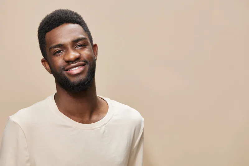Young smiling African male with short hair in a light beige shirt against a neutral background, embodying confidence and positivity