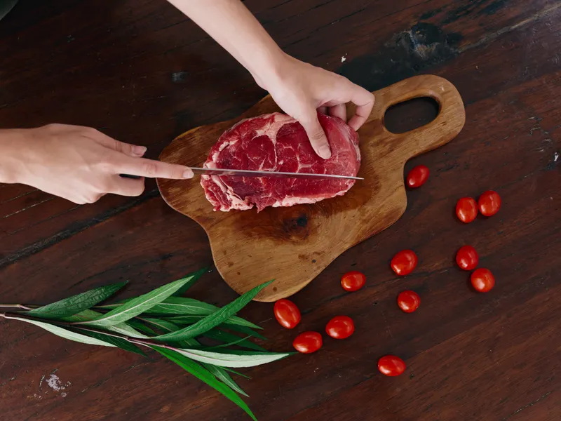 Женщина с ножом в руке нарезает свежее стейковое мясо для жарки на кухне, на столе соль, перец и другие специи, красные помидоры черри и зелень, приготовление ужина