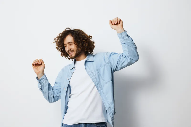 Joyful young man dancing happily in a casual outfit against a light background, expressing carefree emotions and positivity