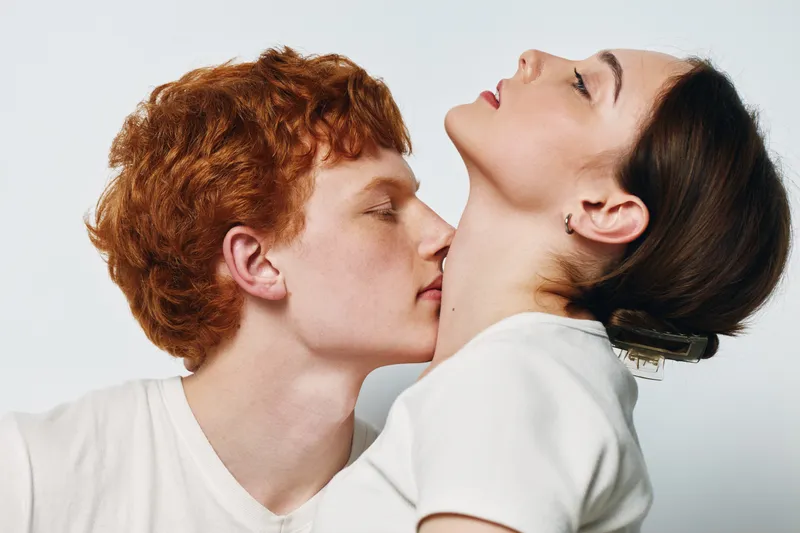 Young man and woman with red hair and brown hair in white shirts showing intimate moment with affection and closeness on light background. Couple with gentle emotion and connection.