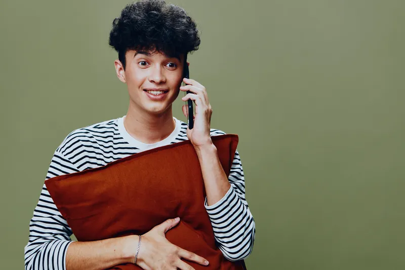 smiling man talking on phone holding orange pillow, wearing striped shirt, casual young adult with curly hair, green background, cheerful expression, indoors
