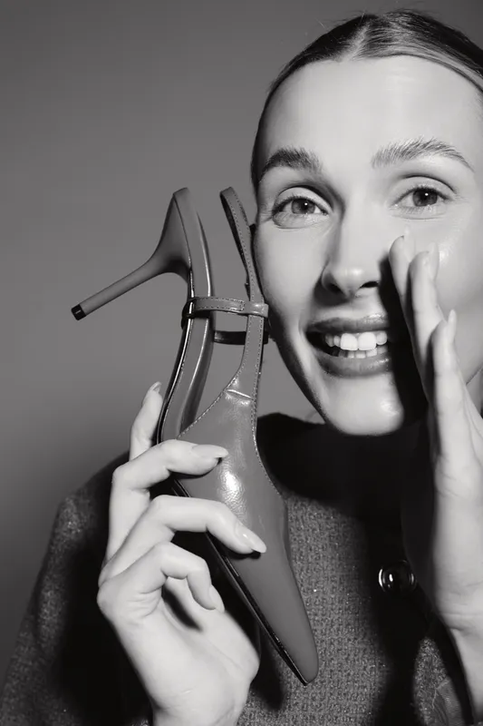 Happy Caucasian woman with fair skin holds high heel shoe near face, smiling and whispering. Studio portrait with monochrome lighting and fashion concept.