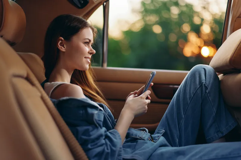 Young woman enjoying a relaxing moment in a car, smiling while using a smartphone, dressed in casual denim, with a soft sunset background.