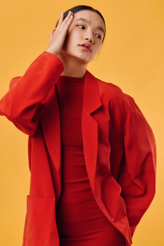 Woman wearing red oversized blazer and dress posing with hand on face, looking sideways against yellow background. Portrait of fashion model in vibrant clothing and contemporary style.