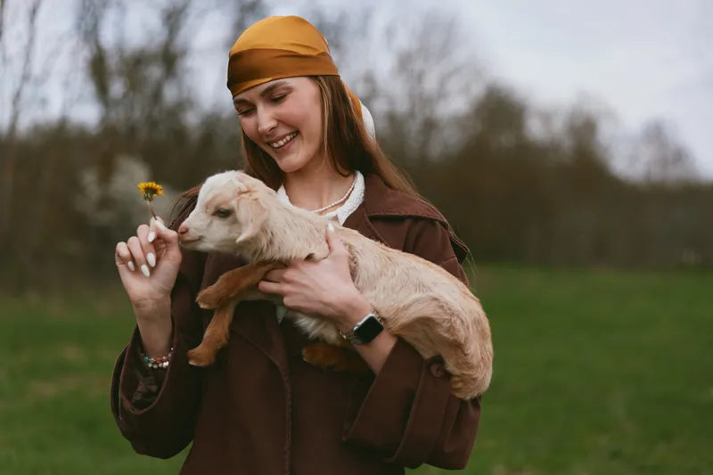 Young woman with long hair and headscarf holds a baby goat and smiles while offering a small flower in a green outdoor field on an overcast day.