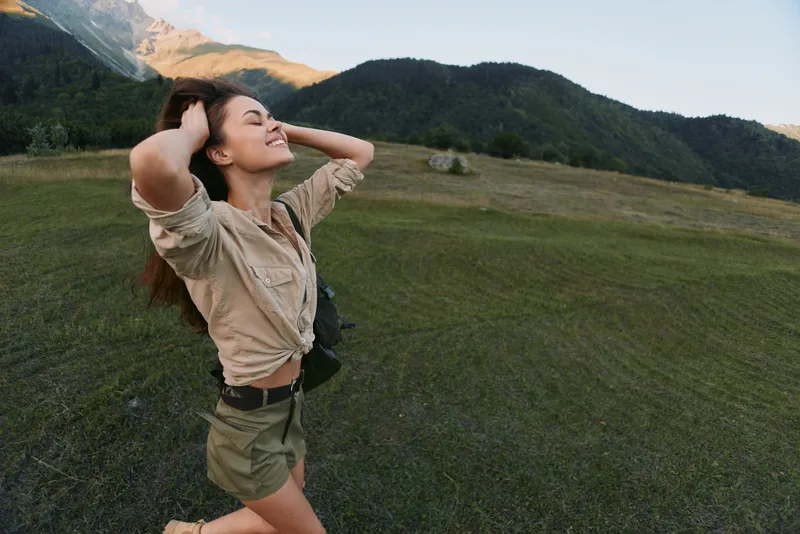 Happy woman in casual clothes enjoying nature with eyes closed and hands in hair. Outdoor lifestyle concept with mountains and green grass in background.