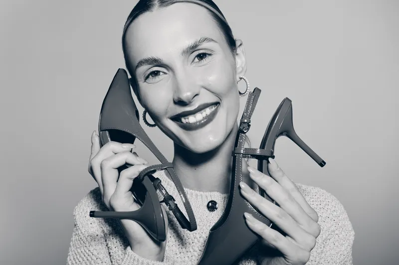 Young Caucasian woman with fair skin smiling and holding two high heel shoes near her face. Concept for fashion, footwear, and happy female model in studio light.
