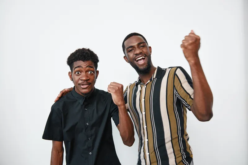 Two young black men with dark skin celebrating success together, smiling and showing excitement in casual clothing against white background in studio portrait.