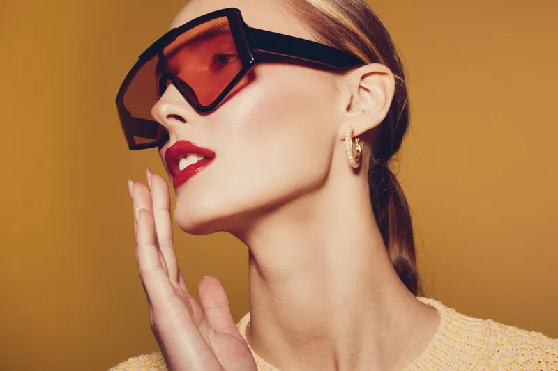 Stylish woman with red lipstick wearing oversized orange sunglasses and gold hoop earrings poses against warm brown background. Fashion concept with modern accessories and makeup.