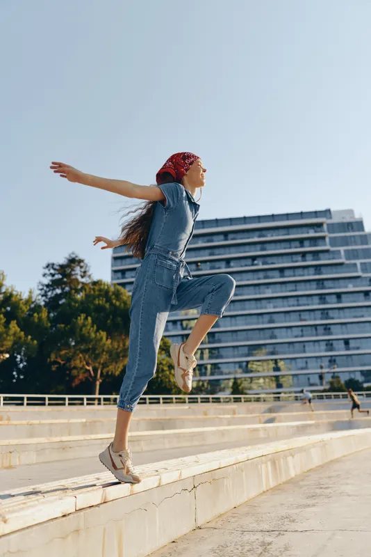 Lifestyle woman in modern boho-western denim street style jumping outdoors, wearing red bandana and casual sneakers under clear blue sky, film color effect.