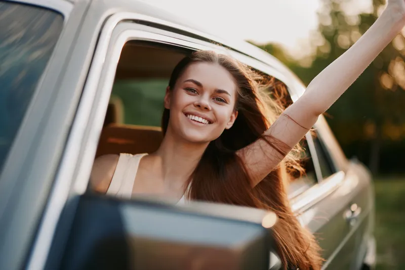 Young woman enjoying a road trip, smiling and waving from a car window, showcasing happiness and freedom in a summer setting with a green landscape.