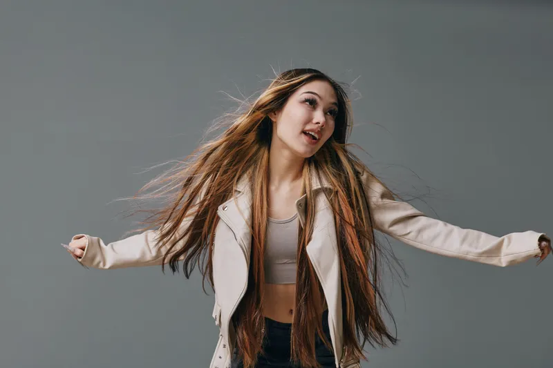 Young woman with long hair is dancing in casual beige jacket and crop top against gray background. Movement, joy and fashion style in studio for lifestyle and youth culture.