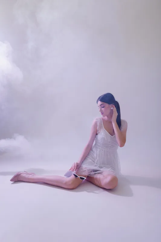 Young woman in white lace dress poses seated on floor with soft lighting and misty background. Concept of beauty, fashion, and elegance in studio setting.