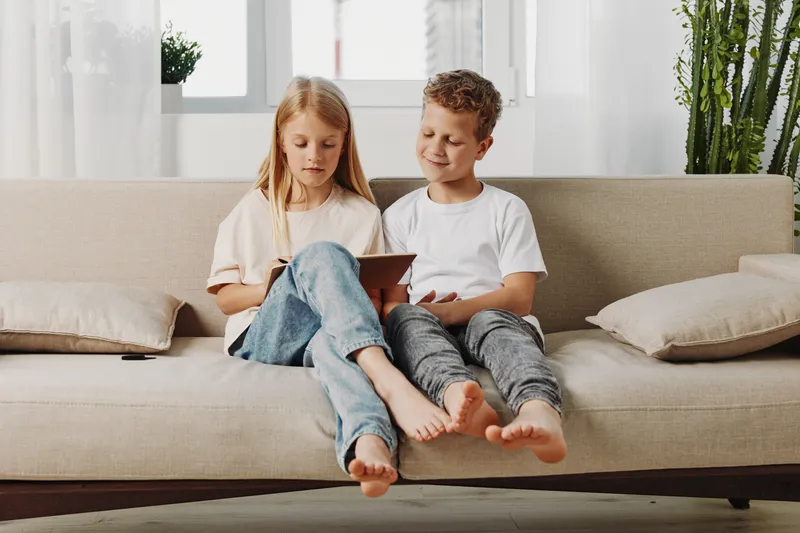 Two barefoot children sitting on a beige sofa in a bright living room, focused on a tablet. Casual clothing and natural light create a cozy, relaxed atmosphere of childhood leisure and technology use.