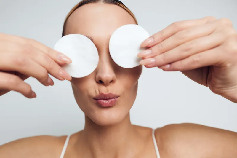 Young woman holding cotton pads over eyes, puckering lips, skincare routine, clean skin, beauty concept, white background, close up portrait, natural light
