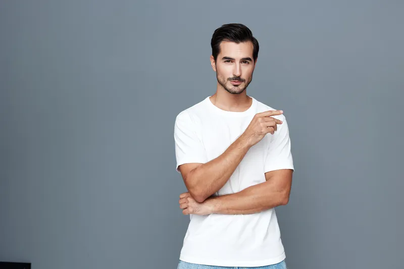 Smiling man wearing a casual white t-shirt stands confidently against a grey background, pointing with one hand. Fashion lifestyle concept.