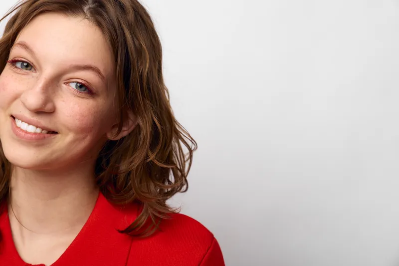 Smiling young woman with brown hair wearing red sweater looks at camera with friendly expression on white background. Concept of happiness, casual style, and natural beauty.