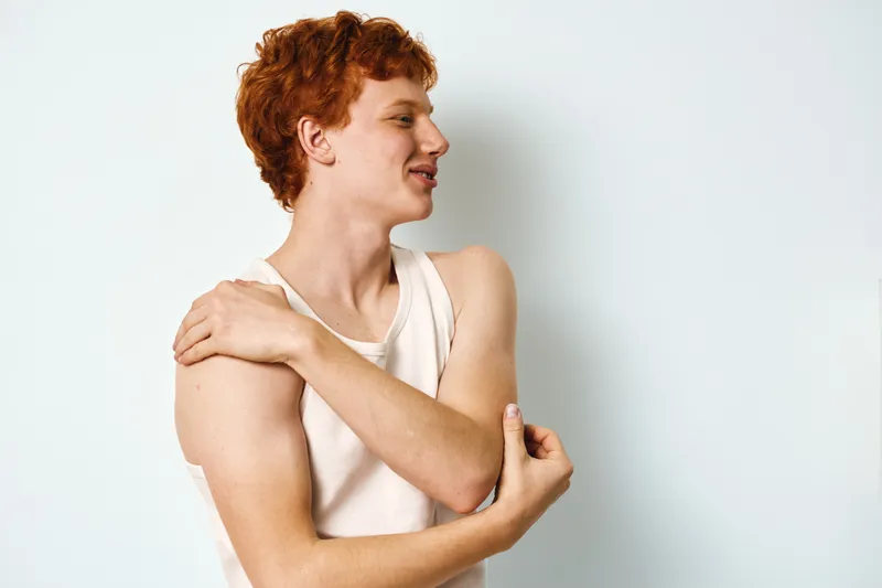 Young man with red hair in white tank top standing against white background, touching shoulder and looking sideways with relaxed and thoughtful expression.