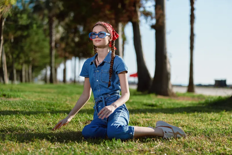 young woman sitting on green grass wearing blue denim outfit and red headscarf outdoors in sunny park with trees and blurred background