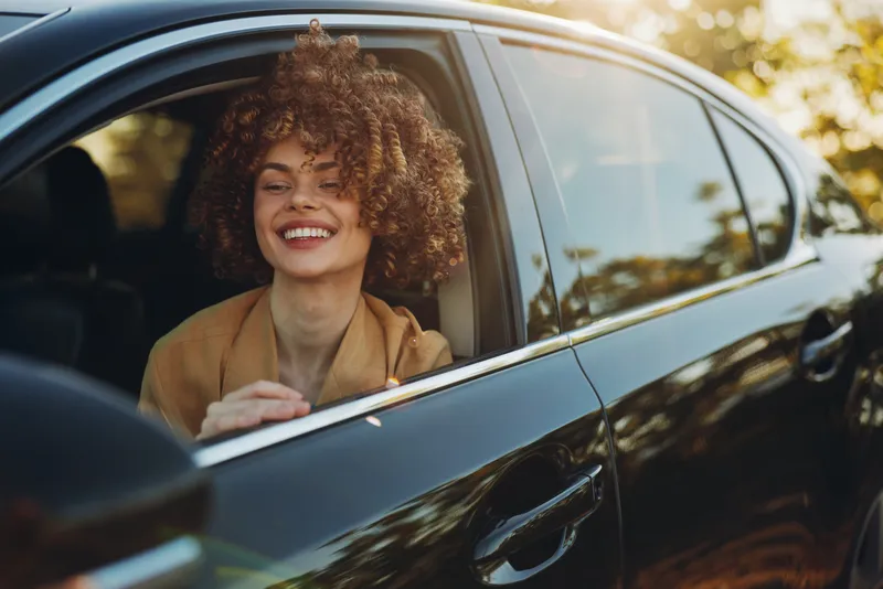 A smiling woman leans from a car window during golden hour, embracing a carefree street style. The retro film color palette adds warmth and nostalgia, capturing everyday joy in motion.
