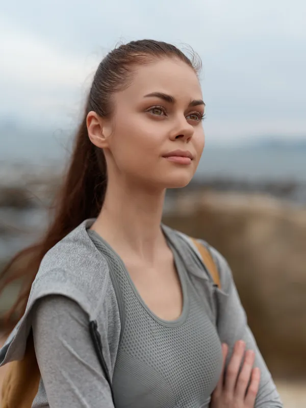 Young woman in sportswear with a confident expression, standing against a serene outdoor backdrop, reflecting a healthy lifestyle and perseverance