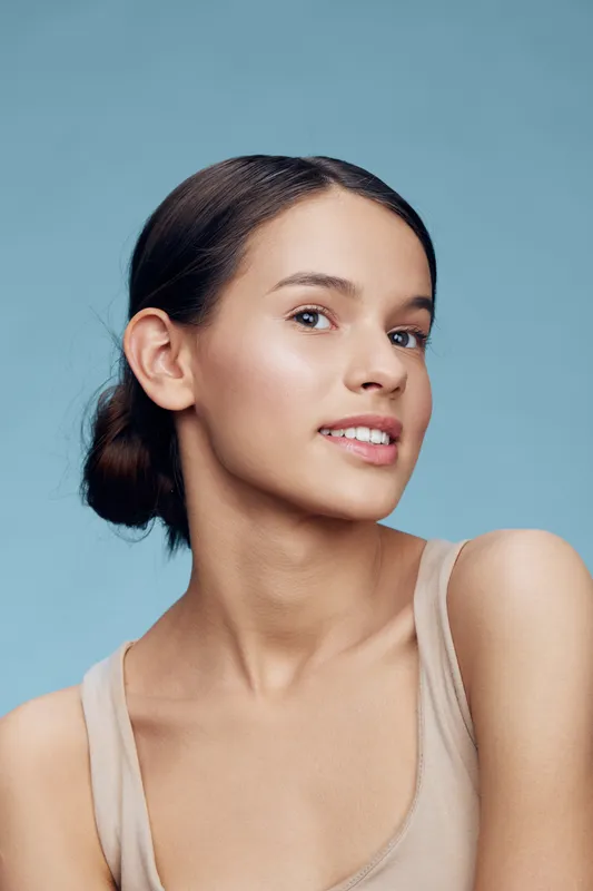Young woman with a radiant smile, natural makeup, and smooth skin, set against a light blue background, capturing elegance and confidence with a trendy style