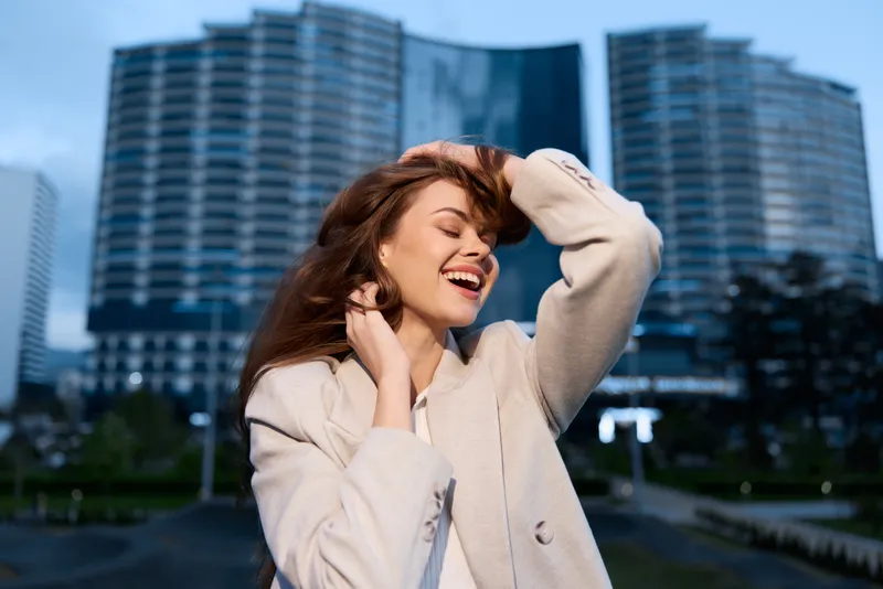 Businesswoman smiling outdoors, dressed in stylish attire, with skyscrapers in the background, conveying confidence and positivity, ideal for corporate use.