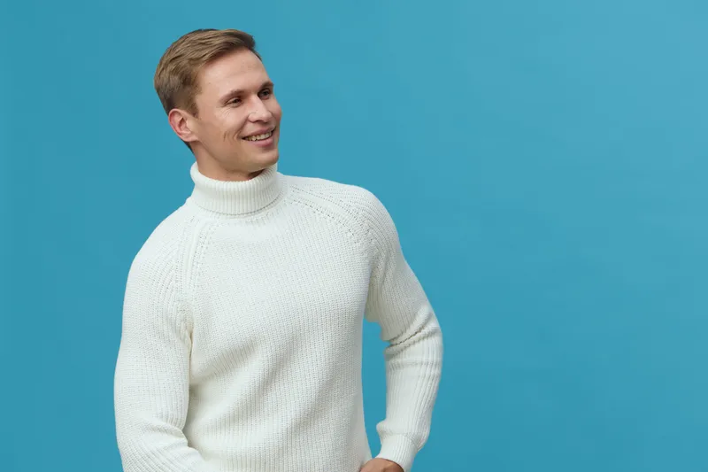 smiling young caucasian man wear white knitted turtleneck sweater look aside isolated on blue background casual style portrait digital nomad concept