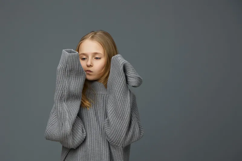 A pensive young girl in an oversized gray sweater, showcasing emotions of contemplation and introspection against a muted gray background